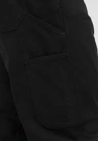Pantaloni cargo neri con una texture liscia, caratterizzati da ampie tasche laterali e un design a pannelli unico. Le cuciture sono visibili lungo i bordi.
