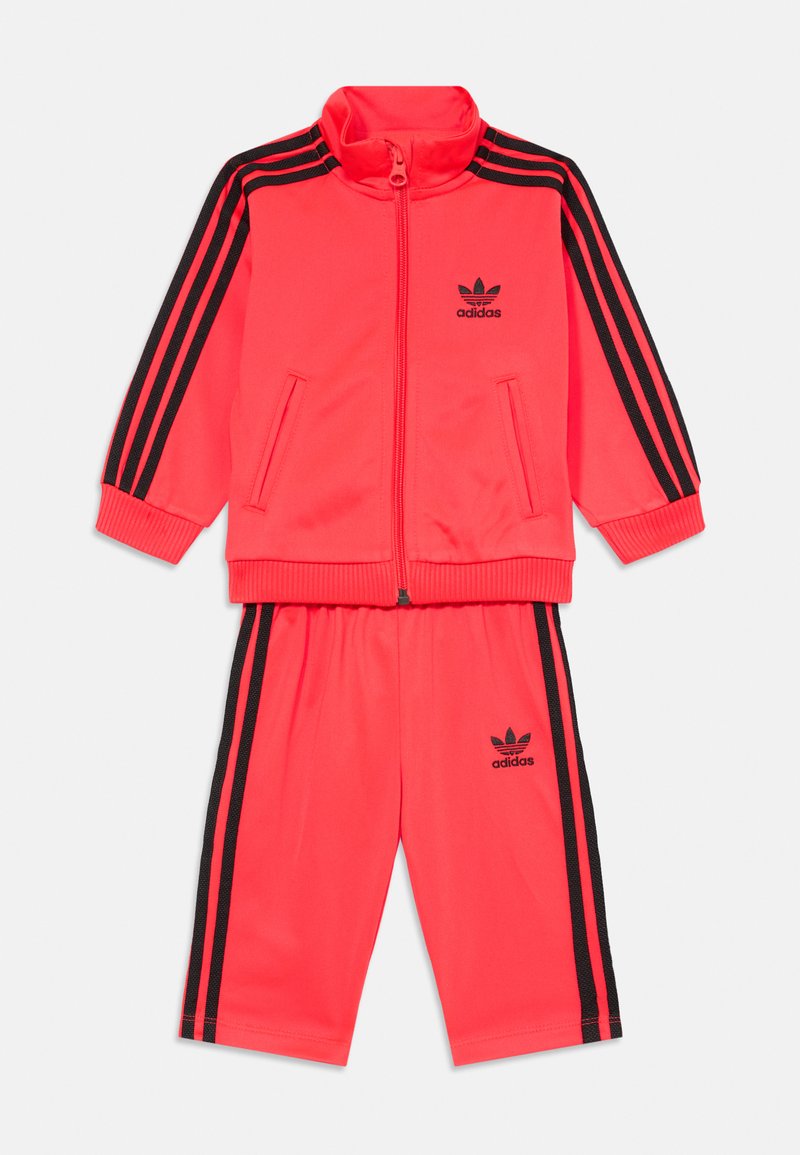 Spilgtas rozā sporta tērpa komplekts, kas sastāv no jaku ar rāvējslēdzēju un augstu apkakli, melnām svītrām un saskaņotām šortiem. Adidas logo abās daļās.