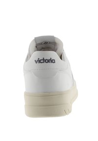 Baskets synthétiques blanches avec une texture lisse, profil bas et semelle en caoutchouc beige. Présente un logo gris brodé au niveau du talon.