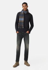 Chemise à manches longues marine, écharpe à carreaux marron et bleu, jean en denim foncé et bottes marron. L'écharpe a des accents à franges et un motif à carreaux.