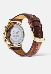 Orologio in acciaio inossidabile oro con cinturino in pelle marrone. Presenta un quadrante circolare, numero di serie visibile e chiusura con fibbia.