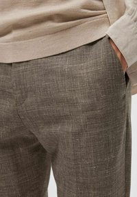 Pantalon en lin marron avec une surface texturée. La taille est partiellement visible, présentant un tricot côtelé en haut.
