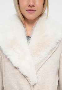 Palton bej cu o cravată mare din blană sintetică albă. Textură moale, material asemănător lânăi, față cu galoane duble și rever tăiat.