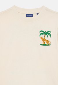 Cremefarbenes T-Shirt mit kurzen Ärmeln, das auf der linken Brustseite einen grünen Palmenbaum und ein gelbgeflecktes Leopardenmotiv zeigt.