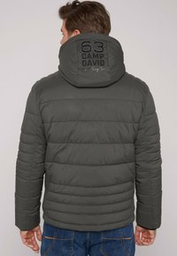 Chaqueta de plumas de color gris oscuro con un alto cuello, textura acanalada y un logotipo prominente en la capucha. Costuras horizontales por toda la prenda.
