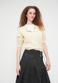 Πλεκτή ζακέτα σε κρεμ χρώμα με σχέδιο διαμαντιού, κουμπωτή μπροστά και ριμπ μανσέτες, συνδυασμένη με σκουρόχρωμη τζιν φούστα με πιέτες και αγκράφες.