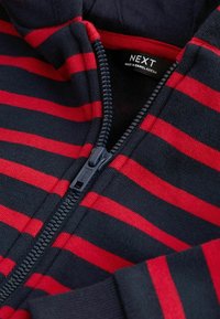 Gros plan sur un vêtement zippé rayé bleu marine et rouge, avec une fermeture éclair partiellement ouverte et une étiquette "NEXT Made in Bangladesh" à l'intérieur du col.