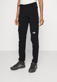 Svarta utomhusbyxor med fickor, tillverkade av lätt material. Har en logotyp på låret och justerbara muddar. Bärs med mörka skor.