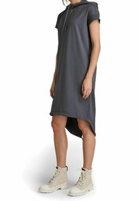 Kvinna i en mörkgrå huvan kortärmad klänning med hög-låg fåll och ljusbeige snörade ankelstövlar.