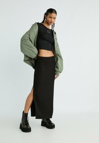 Frau mit zwei geflochtenen Pferdeschwänzen, die eine grüne Daunenjacke, ein schwarzes Crop-Top, einen langen schwarzen Rock mit Seitenschlitz und schwarze Plateau-Stiefel trägt.