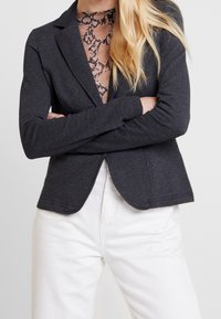 Blazer gris carbón de diseño ajustado, mangas largas y solapas con muesca, superpuesto sobre una blusa con patrón de encaje, combinado con pantalones blancos.