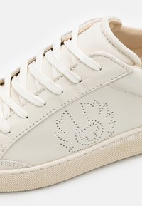 Baskets en cuir blanc avec des lacets plats blancs, un design perforé sur le côté et une semelle en caoutchouc texturée avec un motif diagonal.