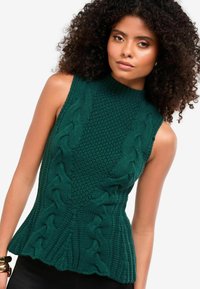 Pulover verde fără mâneci, tricotat, cu guler înalt, având modele texturate tip cablu și o formă peplum, realizat dintr-un fir gros.