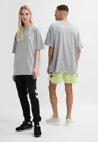 Grå oversized t-shirt med rund halsringning, tillsammans med svarta byxor med vita chevronränder. Den manliga modellen bär limegröna shorts.
