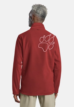 Man met grijs haar draagt een rode fleecejas met een groot wit pootafdrukontwerp op de bovenrug, gecombineerd met beige broek.