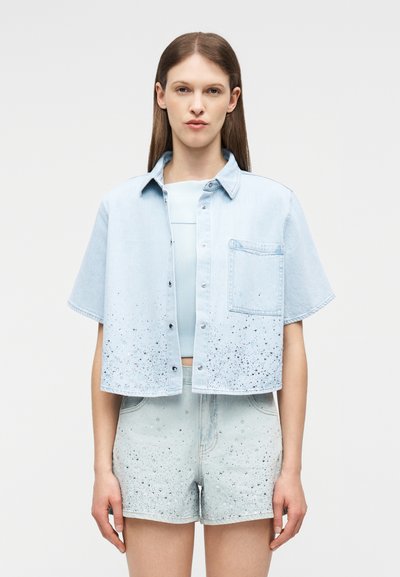 Γυναίκα που φοράει ανοιχτό μπλε τζιν κοντομάνικο πουκάμισο με κουμπιά και μπροστινή τσέπη, καθώς και ταιριαστά κοντά σορτς με ψηλή μέση και σχεδιασμό με στικτές βούλες.
