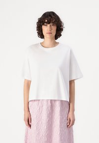 T-shirt blanc à manches courtes avec un col rond, présentant une coupe décontractée. Le modèle porte une jupe rose texturée, mettant en valeur une apparence superposée.