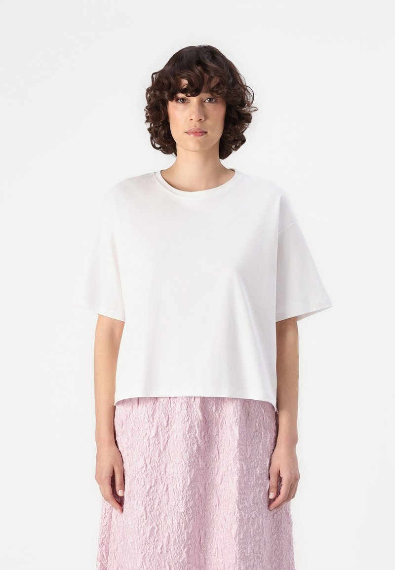 White short-sleeve t-shirt with a round neckline, featuring a relaxed fit. The model wears a textured pink skirt, showcasing a layered appearance.