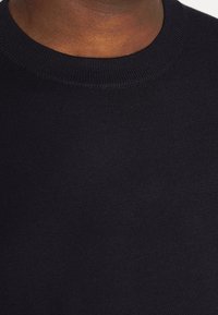 Gros plan sur le cou et le haut du torse d'une personne portant un pull noir uni en tricot avec col rond.