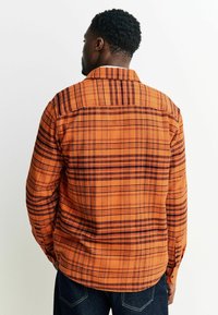 Man in een oranje geruit overhemd met lange mouwen en donkere spijkerbroek, staand met zijn handen achter zijn rug, met zijn gezicht van de camera af.