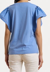 Jasnoniebieska koszulka z krótkim rękawem, z podniesionymi rękawami i dekoracyjnym haftem, wykonana z miękkiej bawełny. Luźny krój, gładki widok z tyłu.