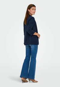Marineblazer med optrukne ærmer, parret med lyseblå flared jeans. Modellen har klare højhælede sandaler på. Glat stof og tætsiddende pasform.