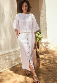 Bílý lněný crop top s širokými rukávy, který je kombinován s bílou midi sukní na knoflíky s bočním rozparkem a béžovou tkanou taškou. Ploché sandály.