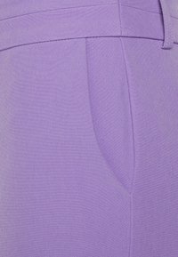 Närbild av en lavendelfärgad tygplagg som visar en sidficka och bältesöglor med detaljerad sömnad.