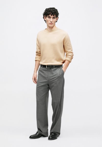 Jeune homme aux cheveux bouclés portant un pull beige, un pantalon plissé gris, une ceinture noire et des chaussures habillées noires, debout avec une main dans la poche.