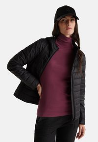 Giubbotto puffo nero con texture trapuntata, indossato su un dolcevita bordeaux, abbinato a dei jeans neri. Un cappellino nero completa l'outfit.