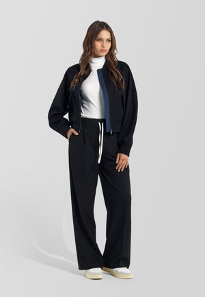 Mujer con chaqueta bomber negra sobre un jersey de cuello alto blanco, pantalones negros de pierna ancha con cordón blanco y zapatillas blancas, de pie.