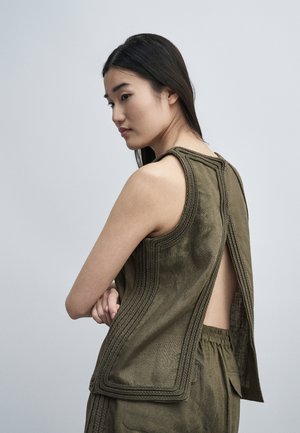Femme aux longs cheveux foncés portant un haut olive sans manches avec un dos ouvert et un pantalon assorti, debout devant un fond gris uni.