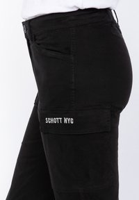 Zwarte cargo broek met een zijzak waarin "SCHOTT NYC" in het wit geborduurd is. De stof is soepel, met een slanke pasvorm en knoopsluiting.