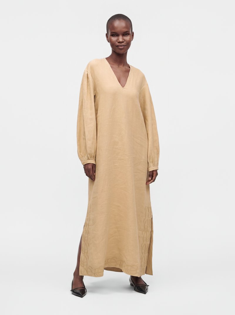 Femme noire debout dans une longue robe beige avec un col en V, des manches longues, des fentes latérales et des chaussures noires pointues sur un fond blanc uni.