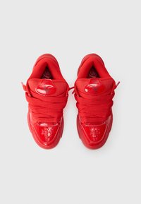 Scarpe da ginnastica rosse brillanti con lacci spessi viste dall'alto su uno sfondo grigio, dotate di dettagli lucidi e di un logo ricamato sulla linguetta.