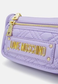 Fialová taška z umělé kůže s prošívanou texturou, zlatými prvky a výraznou destičkou s logem "LOVE MOSCHINO" na přední straně.