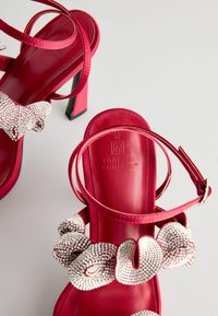 Rote High Heels mit satinierter Oberfläche, ausgestattet mit einem mit Strasssteinchen verzierten Riemen und Fesselverschluss, vor einem hellgrauen Hintergrund.