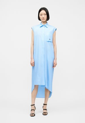 Žena v svetlomodrých midi šatách bez rukávov s asymetrickým lemom a čiernymi sandálmi na vysokom opätku s remienkami.