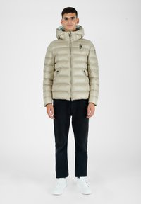Beige pufjakke med quiltet design, lynlås-lukning og hætte. Båret over sorte bukser og hvide sneakers, med sidelommer.