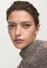 Jeune femme aux cheveux courts et foncés, portant un pull en tricot marron à col haut, regardant directement l'appareil photo devant un fond clair.