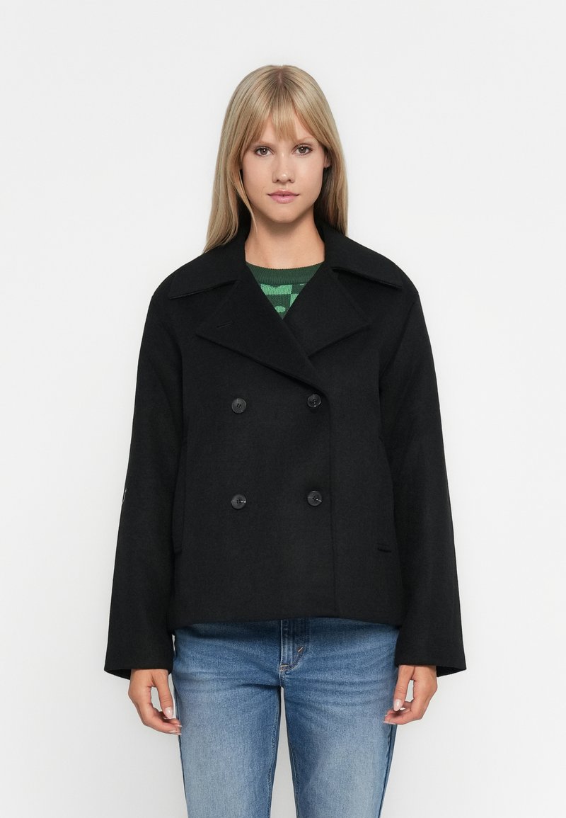 Svarte dobbeltradte peacoat med bred krage, utstyrt med store svarte knapper og en firkantet passform, kombinert med en grønnstripete genser.