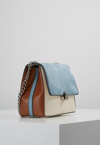 Kabelka se strukturovaným designem, s modrým semišovým klopou, krémovým tělem a hnědou stranou. Obsahuje stříbrné kovové prvky a řetězový popruh.