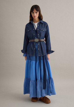 Frau trägt ein dunkles Jeanshemd über einem weißen Oberteil, mit Gürtel in der Taille, dazu einen mehrstufigen, blau gestreiften Maxirock und braune Mokassin-Schuhe.