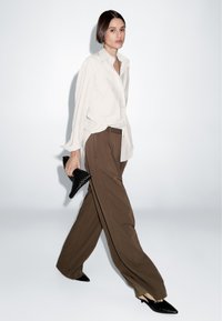 Vrouw die loopt in een witte oversized blouse, brown wijde broek, zwarte puntige hakken, en een zwarte gestructureerde clutch draagt.