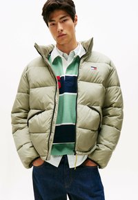 Chaqueta acolchada de color verde claro, con cierre de cremallera y bolsillos. Llevada sobre una camisa a rayas verde y azul marino, con jeans oscuros.