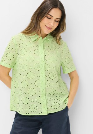 Femme portant une chemise à manches courtes vert clair avec des motifs découpés circulaires, associée à un pantalon bleu foncé, debout, les mains dans les poches.
