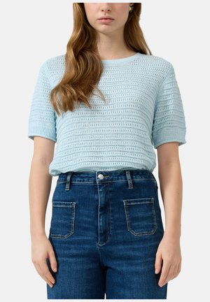 Kobieta w jasnoniebieskim, krótkim sweterku z poziomym wzorem ażurowym i ciemnoniebieskich, wysokich spodniach dżinsowych z przednimi naszywanymi kieszeniami.