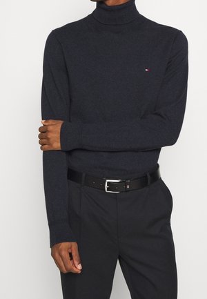 Homme portant un pull à col roulé gris foncé ajusté avec un petit logo sur la poitrine, un pantalon noir et une ceinture noire avec une boucle argentée.