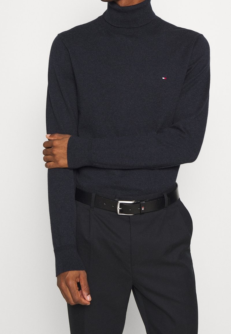 Homme portant un pull à col roulé gris foncé ajusté avec un petit logo sur la poitrine, un pantalon noir et une ceinture noire avec une boucle argentée.