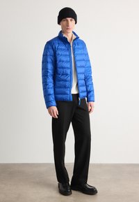Blå pufferjakke med quiltet design, høj krave, frontlynlås og sort hue. Båret med en cremefarvet sweater og sorte bukser.